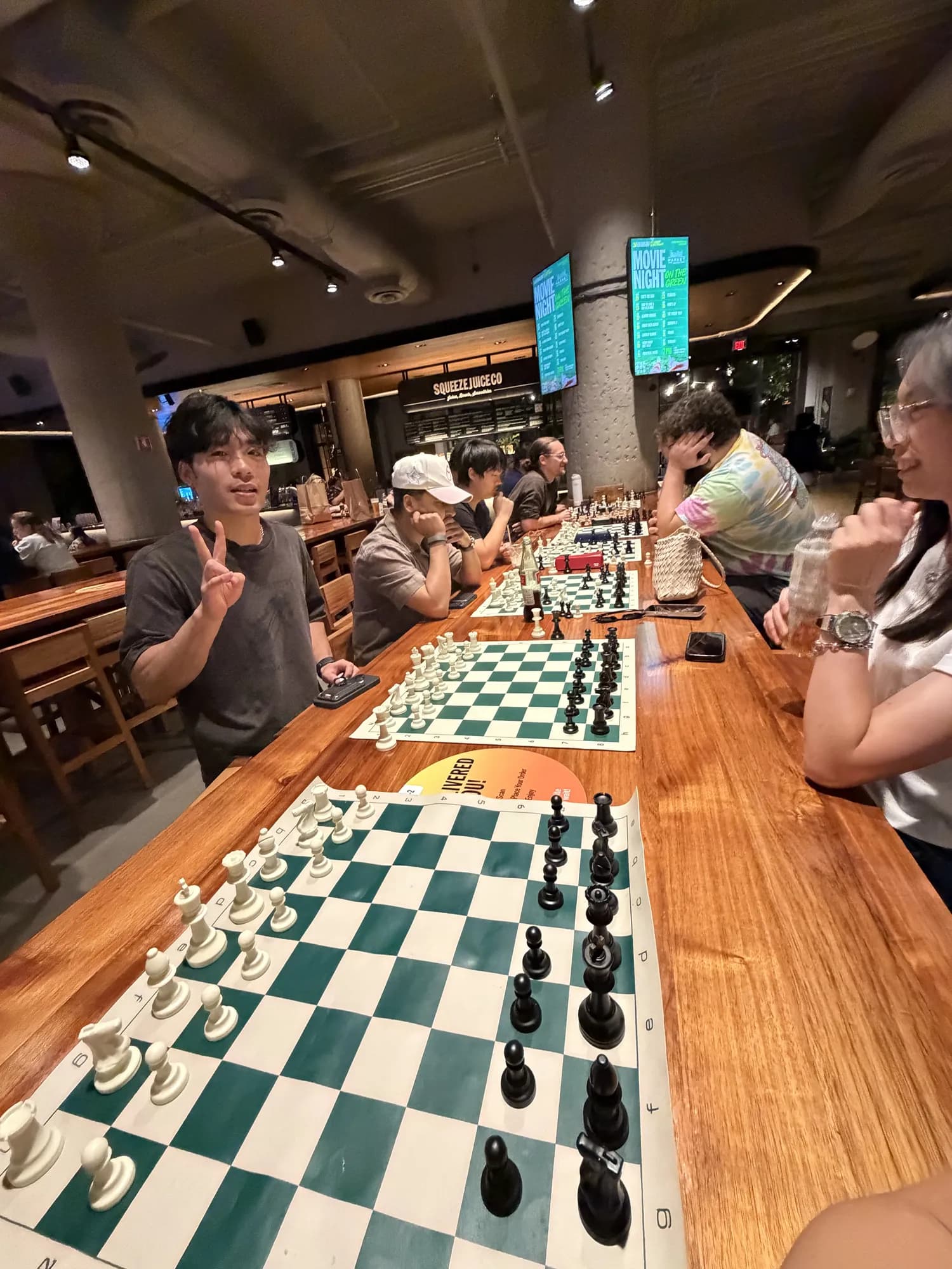 A player flashes a peace sign beside a row of chess boards on a long wooden table