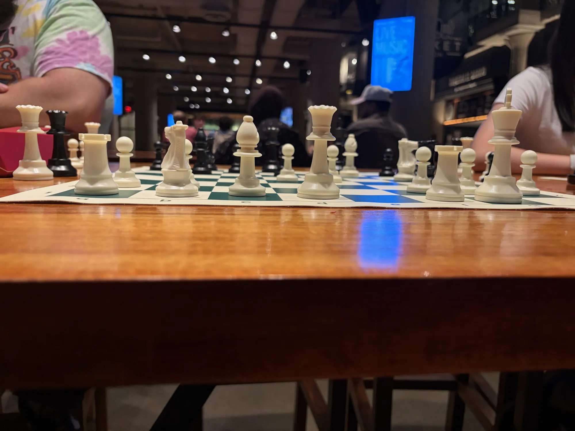 Close-up of chess pieces lined up on a board, with the food hall blurred behind