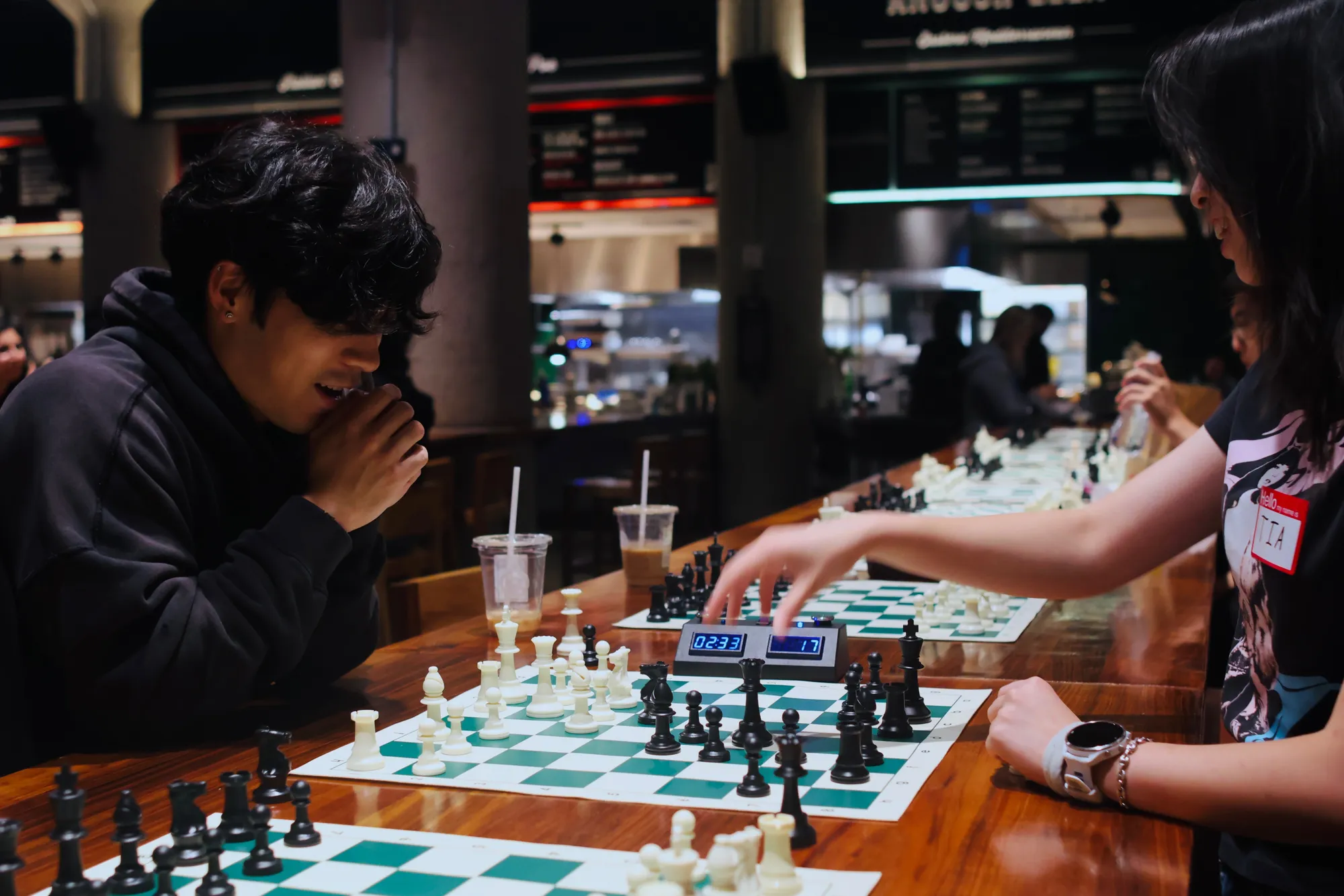 Ed (left) and Tia (right) playing each other at Time Out Market. Tia reaches across the board toward the chess clock while Ed thinks.